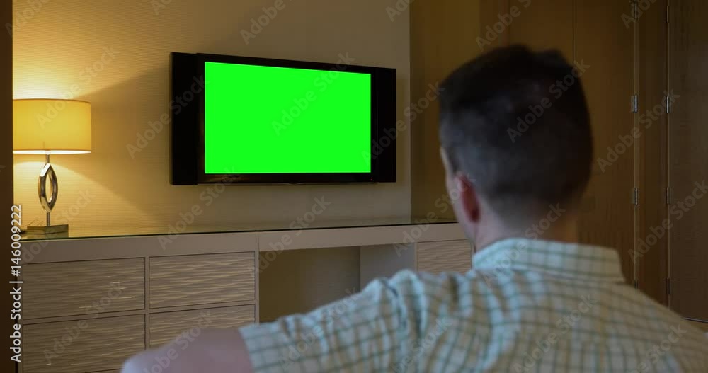 A man watches a television in a luxury hotel room. From behind, with green screen for customizable content. A man watches a television in a luxury hotel room. From behind, with green screen for customizable content.