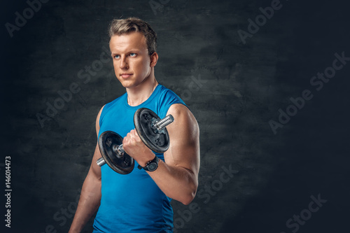 A man dressed in a blue sportswear holds the dumbbell over dark grey background. © Fxquadro