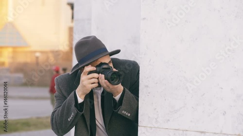 Young male spy agent wearing hat and coat photographing criminal people and hiding behind the wall photo