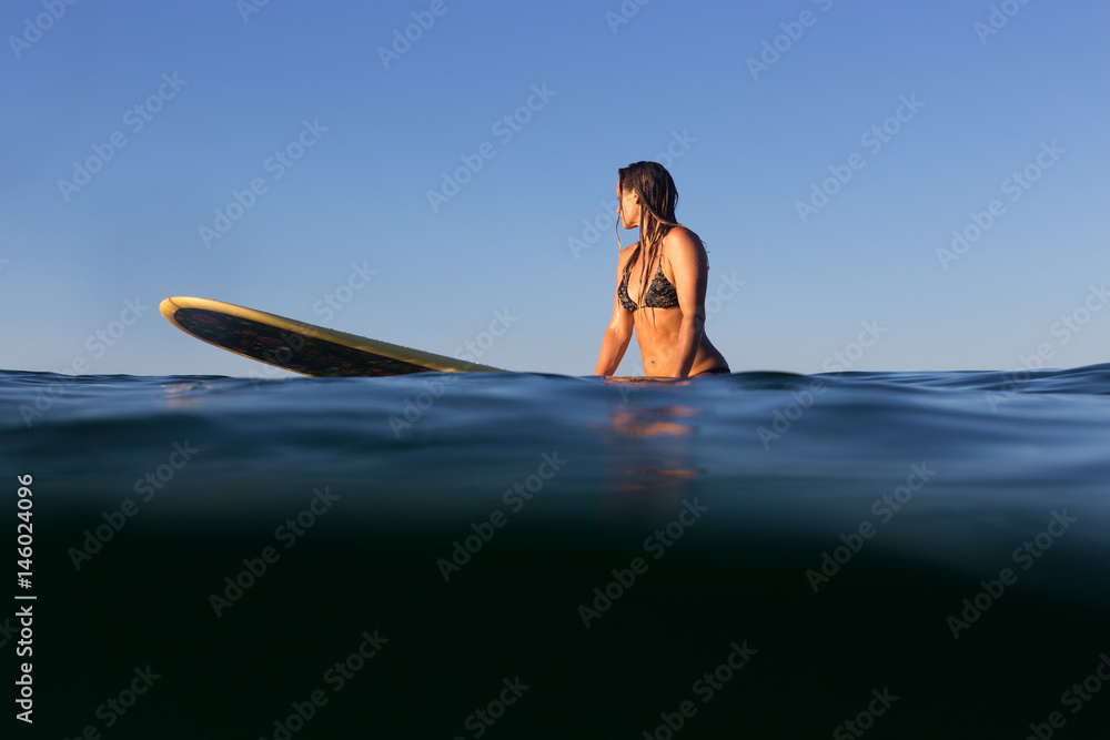 A beautiful female surfer sits on her surfboard in the evening light on the Pacific Ocean.