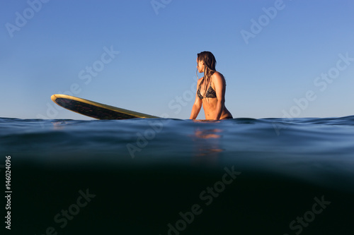 A beautiful female surfer sits on her surfboard in the evening light on the Pacific Ocean.