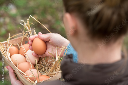 a young farmer female holding a fresh hen egg and other eggs in a basket with nest straw, soft focus photo