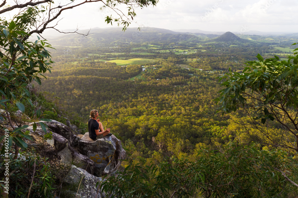 A young man sits, quietly meditating on top of a mountain with expansive views near Noosa Heads, Australia.