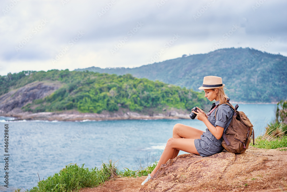 Photo shooting and traveling. Young woman in hat with rucksack holding camera enjoying sea view.