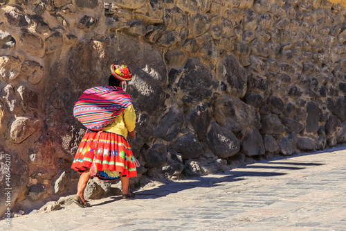 Peruvian woman cholita dressed in traditional colorful cloth, carrying the sack and walking up the street with stony walls, Inkan Sacred Valley, Ollantaytambo, Peru photo