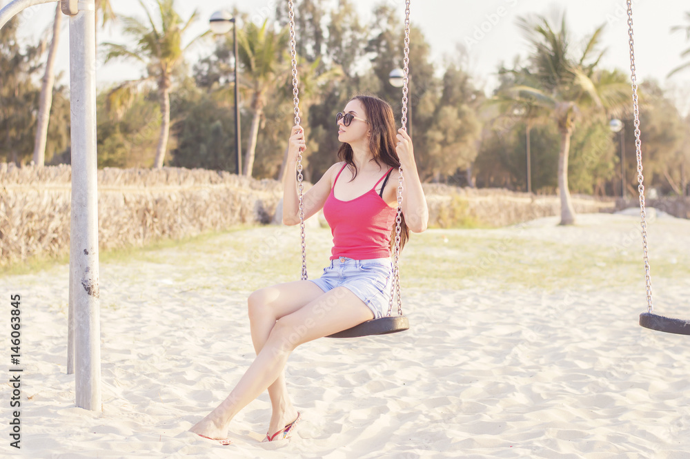 Young european female swinging at Dubai beach. Brunette with long hair in pink top and denim short relaxing at the beach. Vacation concept. Leisure and lifestyle