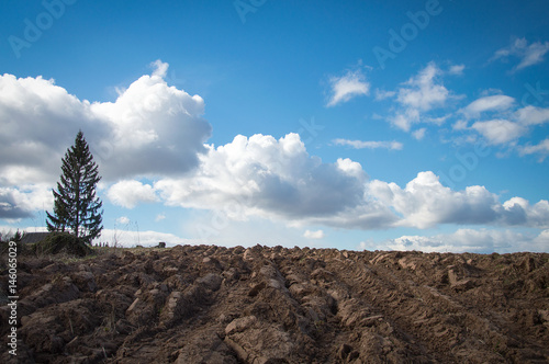 Freshly plowed heavy clay soil in early spring contrasting with blue sky and puffy white clouds. A single tree at the side of the big field.
