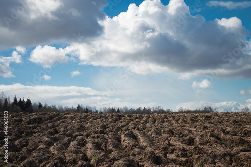 Big field of freshly plowed heavy clay soil in early spring contrasting with blue sky and puffy white clouds. Silhouettes of trees and a forest in the distance.