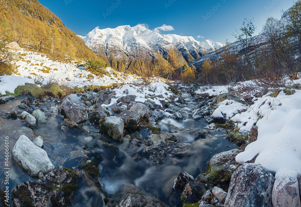 Mountain river flow among rocks; rapid water, golden trees, snow-covered mountains - autumn in Norway.