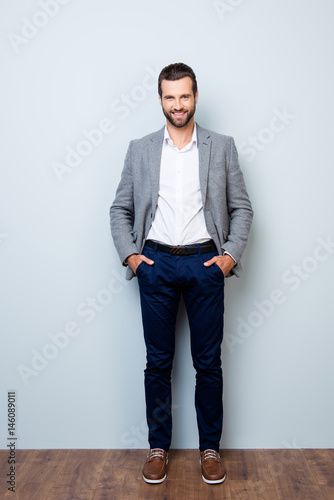 Vertical full-length portrait of handsome smiling happy successful businessman holding his hands in pockets on gray background