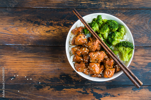 Chicken teriyaki, rice and broccoli in a white big bowl with chopsticks above. Wooden rustic table, top view.