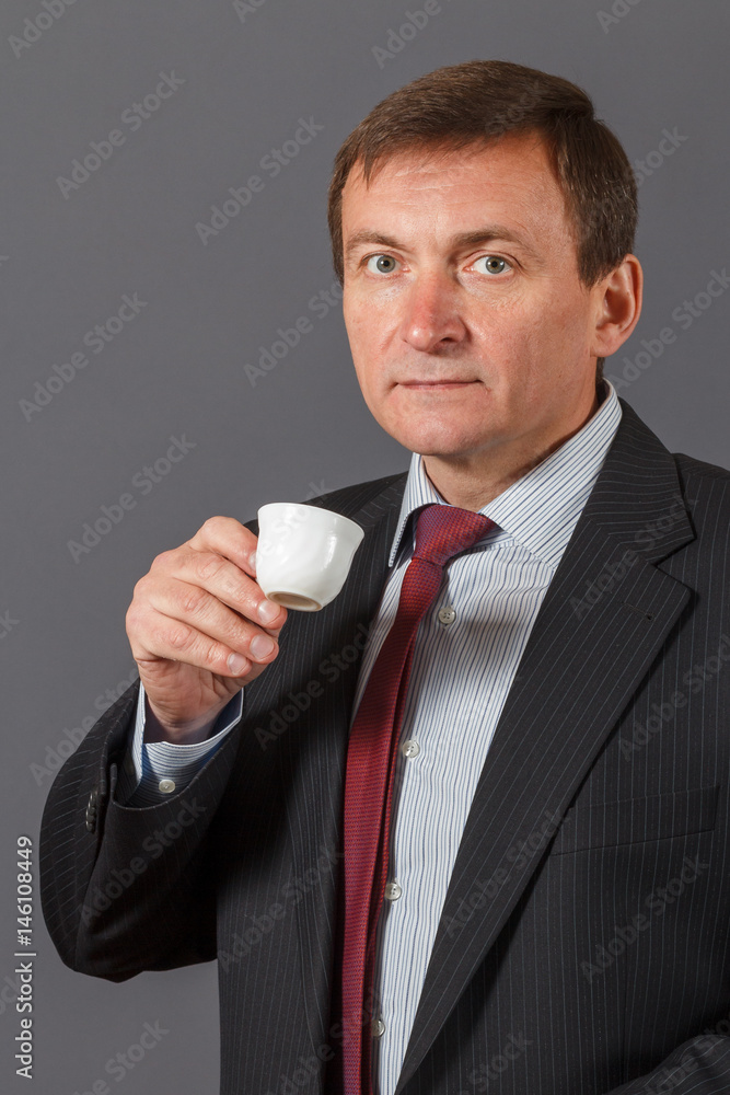 Confident mature businessman standing in front of a grey background taking a cup of coffee Confident mature businessman standing in front of a grey background taking a cup of coffee