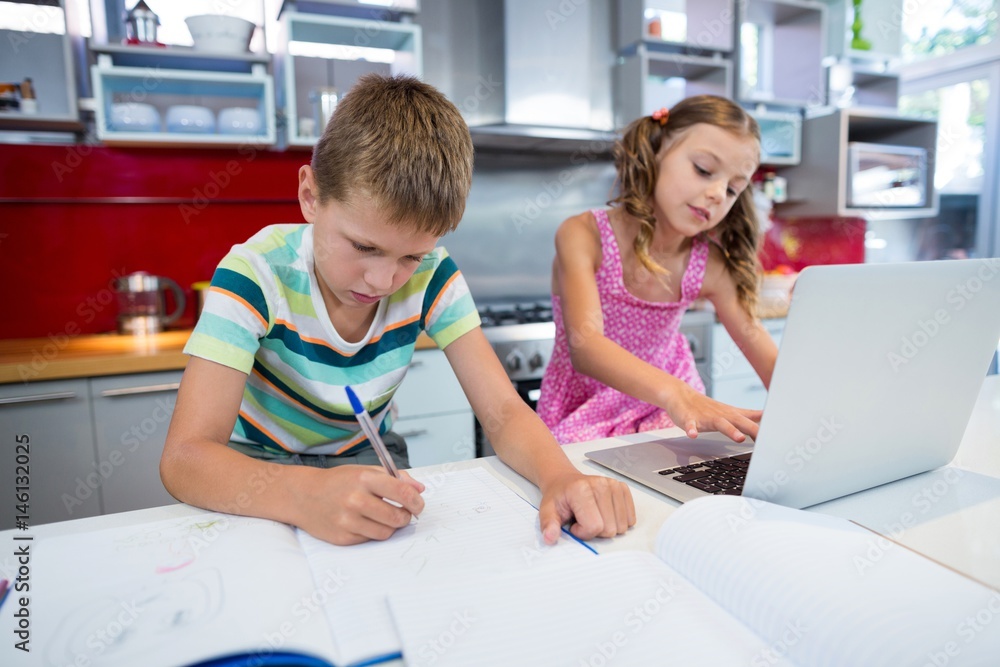 Boy doing his homework while girl using laptop in kitchen Boy doing his homework while girl using laptop in kitchen
