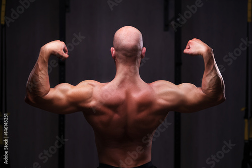 Young healthy bald ripped man with big muscles posing in gym, view from the back