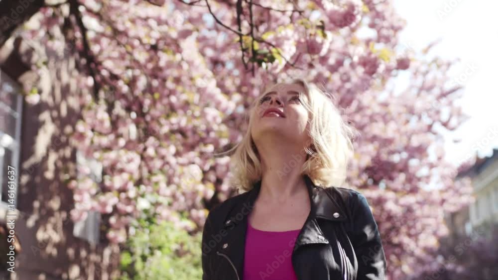 Attractive blue-eyed girl in a casual wear walking down the sakura alley, touching her hair, dreamily smiling, enjoying the blossom, looking to the camera. Modern fashion, positive emotions. Attractive blue-eyed girl in a casual wear walking down the sakura alley, touching her hair, dreamily smiling, enjoying the blossom, looking to the camera. Modern fashion, positive emotions.