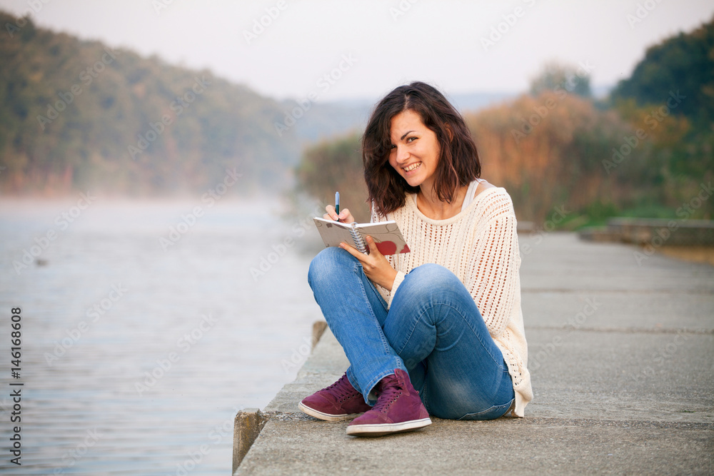 Obraz premium Writing in a notebook. Happy woman is writing her diary in the morning. Sitting by lake.