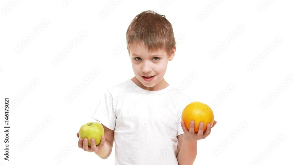 Smiling Caucasian elementary-school aged boy standing on white background with apple and orange in hands and smelling them in slowmotion Smiling Caucasian elementary-school aged boy standing on white background with apple and orange in hands and smelling them in slowmotion