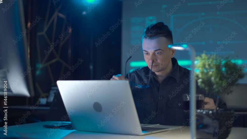 Tyred programmer or Developer sitting at laptop having coffee in modern office late at night Tyred programmer or Developer sitting at laptop having coffee in modern office late at night