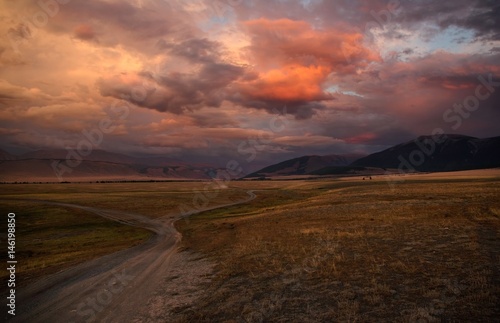 Road path on a desert wild mountain plateau at the background of the hills under a dramatic sunset colorful sky with illuminated red pink purple clouds Kurai Altai Mountains Siberia Russia