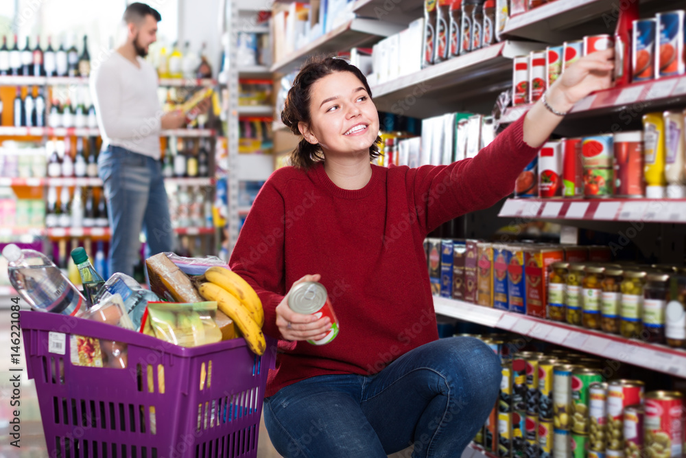 positive young couple choosing purchasing canned food for week at supermarket