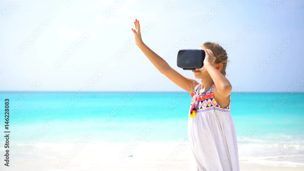 Cute little child girl using VR virtual reality goggles. Adorable girl look into the virtual glasses on white beach Cute little child girl using VR virtual reality goggles. Adorable girl look into the virtual glasses on white beach