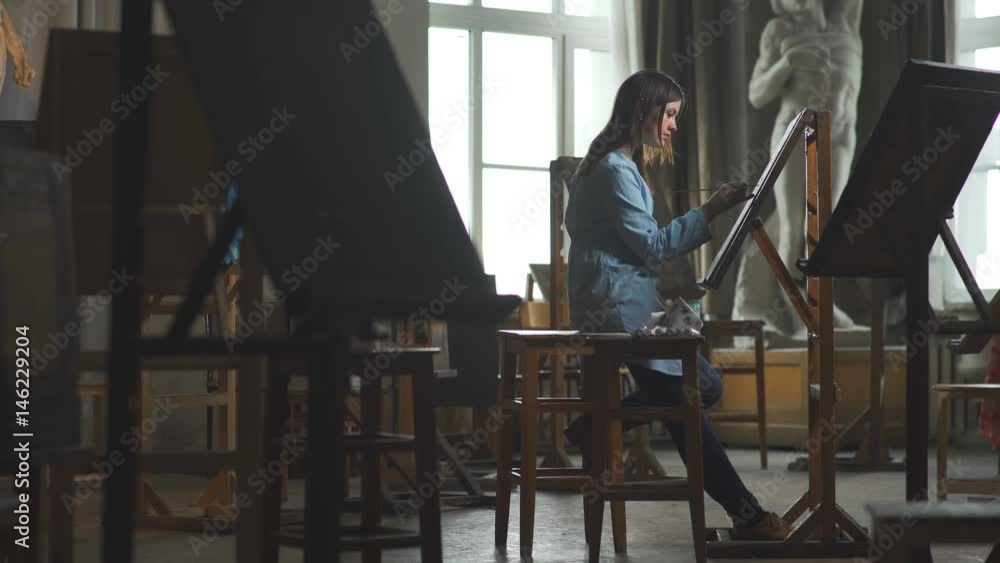 Young woman painter in blue dress draws behind an easel in a light and spacious art studio.General plan: the artist's workshop with interesting creative objects of the interior. Young woman painter in blue dress draws behind an easel in a light and spacious art studio.General plan: the artist's workshop with interesting creative objects of the interior.