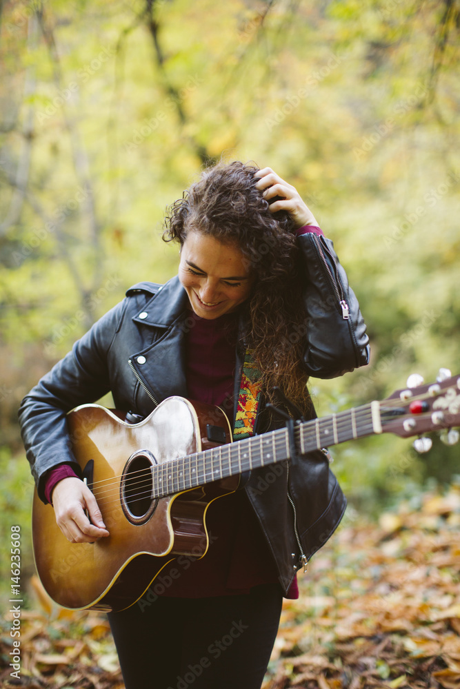 Portrait of beautiful smiling woman playing guitar on forest, fashion lifestyle. Girl wearing black jacket.