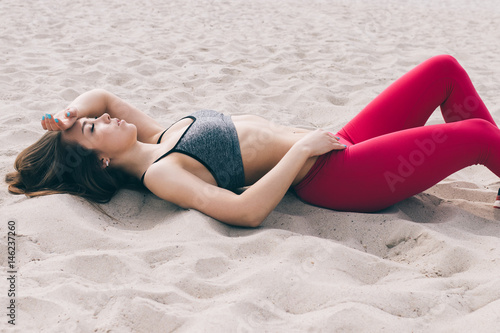 Beautiful young girl in a sports bra and red pants lies on the beach sand