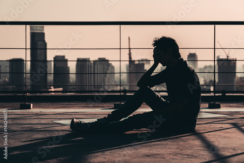 Silhouette of sad depressed Asian man lost hope and cry, sit on building rooftop at sunset, dark mood tone. Concept of major depressive disorder, friend zone, unemployment, stress emotion or paranoid photo