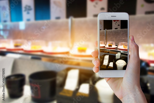 Hand holding smartphone with blur of conveyor belt sushi and sunshine background.