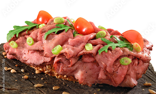 Thinly sliced rare roast beef open sandwich on wholegrain bread. Rocket, tomato and spring onion garnish.