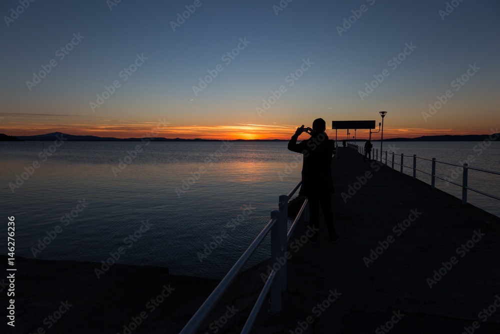 A backlit silhouette of a man on a pier taking a photo of the sunset with his phone, with some other people in the background A backlit silhouette of a man on a pier taking a photo of the sunset with his phone, with some other people in the background