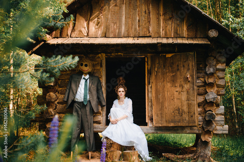 Young beautiful skinny bride girl in vintage wedding dress and hairstyle standing beyond old wooden house in forest with scarecrow from fairytale. Classical russian fable concept and idea. Sorcery. photo