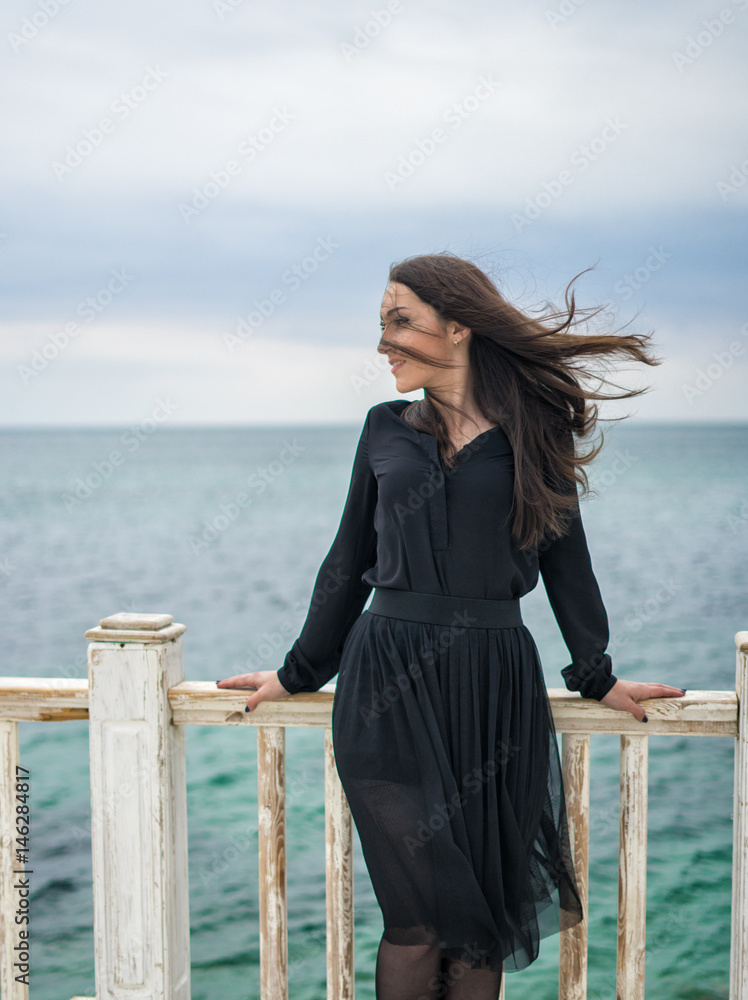 Emotional beautiful brunette girl in a black dress posing on the pier. Sea and grey clouds on background. The wind is blowing her hair. Emotional beautiful brunette girl in a black dress posing on the pier. Sea and grey clouds on background. The wind is blowing her hair.