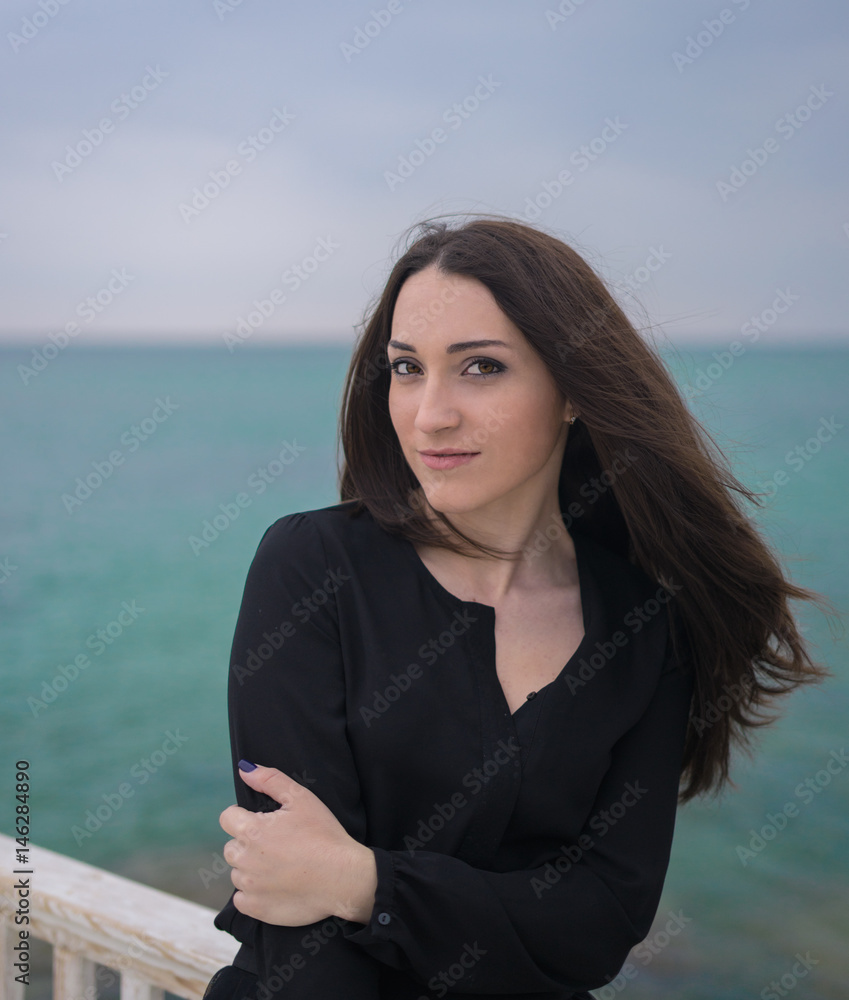 Portrait of Emotional beautiful brunette girl in a black dress posing on the pier. Sea and grey clouds on background. Portrait of Emotional beautiful brunette girl in a black dress posing on the pier. Sea and grey clouds on background.