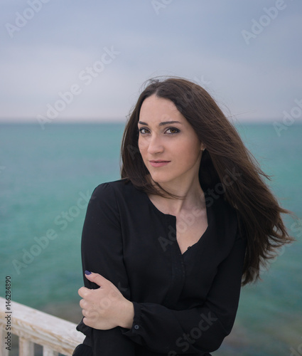 Portrait of Emotional beautiful brunette girl in a black dress posing on the pier. Sea and grey clouds on background. © Igor Kardasov