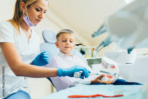 Pediatric female dentist educating a smiling little boy about proper tooth-brushing, demonstrating on a model. Early prevention, raising awareness, oral hygiene demonstration concept.