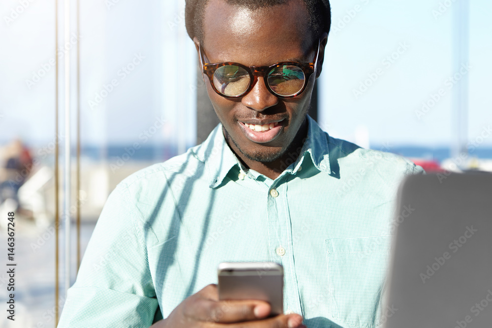 Modern technologies, online communication and people concept. Joyful fashionable young Afro American hipster in shirt and eyewear using mobile phone at airport waiting for girlfriend from trip