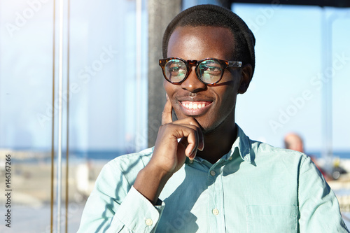 Headshot of cheerful young dark-skinned man wearing trendy glasses and hat touching his face and smiling broadly while thinking about something pleasant, sitting against blurred window background