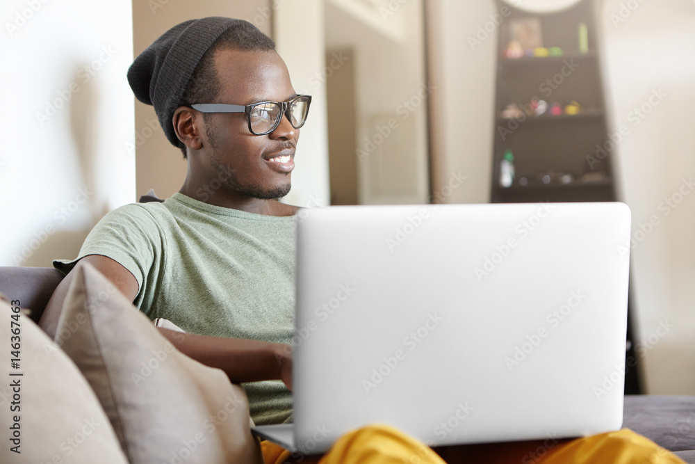 Carefree and relaxed young dark-skinned student in hat and glasses relaxing at home after college, sitting on couch with laptop pc on his lap, playing video games or watching favourite series online Carefree and relaxed young dark-skinned student in hat and glasses relaxing at home after college, sitting on couch with laptop pc on his lap, playing video games or watching favourite series online
