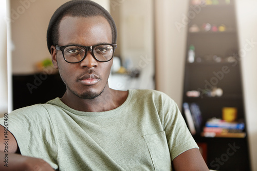 Indoor shot of attractive young African American man wearing trendy black glasses and hat spending weekend morning at home, sitting in living room, watching world news on TV, having serious look