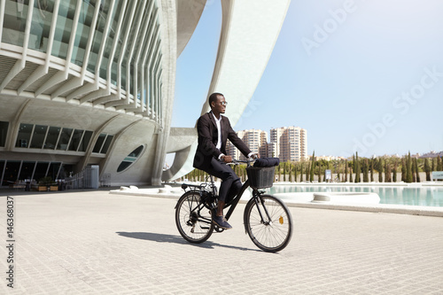 Fototapeta Naklejka Na Ścianę i Meble -  Confident eco friendly dark-skinned CEO using two wheeled pedal assist vehicle to get to work. Successful modern black businessman riding bicycle to office after lunch . People, transport and business