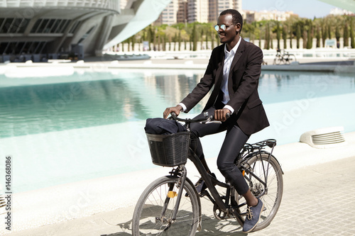 Environmentally friendly Afro American banker in formal wear and shades looking happy and relaxed, cycling to work on bike in urban setting, smiling cheerfully. Businesspeople, lifestyle and transport © wayhome.studio 