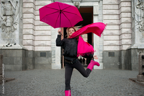 Beautiful young and happy blond woman with colorful umbrella on the street. Girl in a bright pink scarf, rubber boots and umbrella walking in a rainy city.