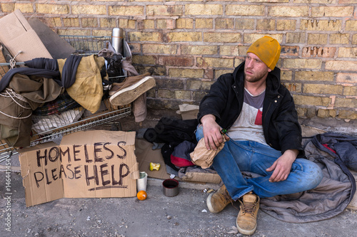 Homeless man holding bottle of alcohol. Sitiing near the cart with his stuff on the street. photo