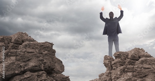 Business man hands in air on rocks against clouds with flare