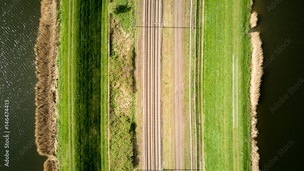 Fototapeta premium Train lines. Vertical aerial drone view of a pair of train tracks bordered by water and pathways creating abstract lined textures.