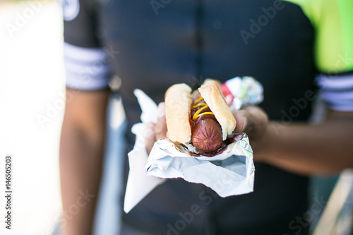 cyclist makes a stop for lunch of huge hot dog with white bun and red ketchup yellow mustard as center focus of photo.