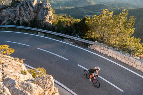Young female cyclist riding, cycling on sunset ray glissened road high in the spainish mountians narrow road surrounded by green trees and huge sand colored rocks.
