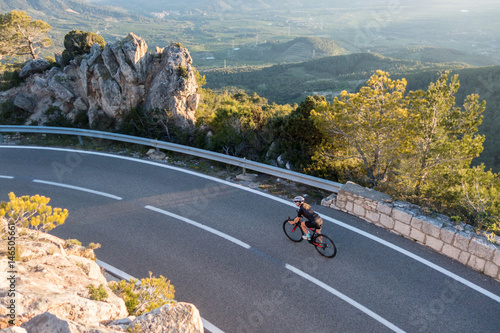 Young female cyclist riding, cycling on sunset ray glissened road high in the spainish mountians narrow road surrounded by green trees and huge sand colored rocks.